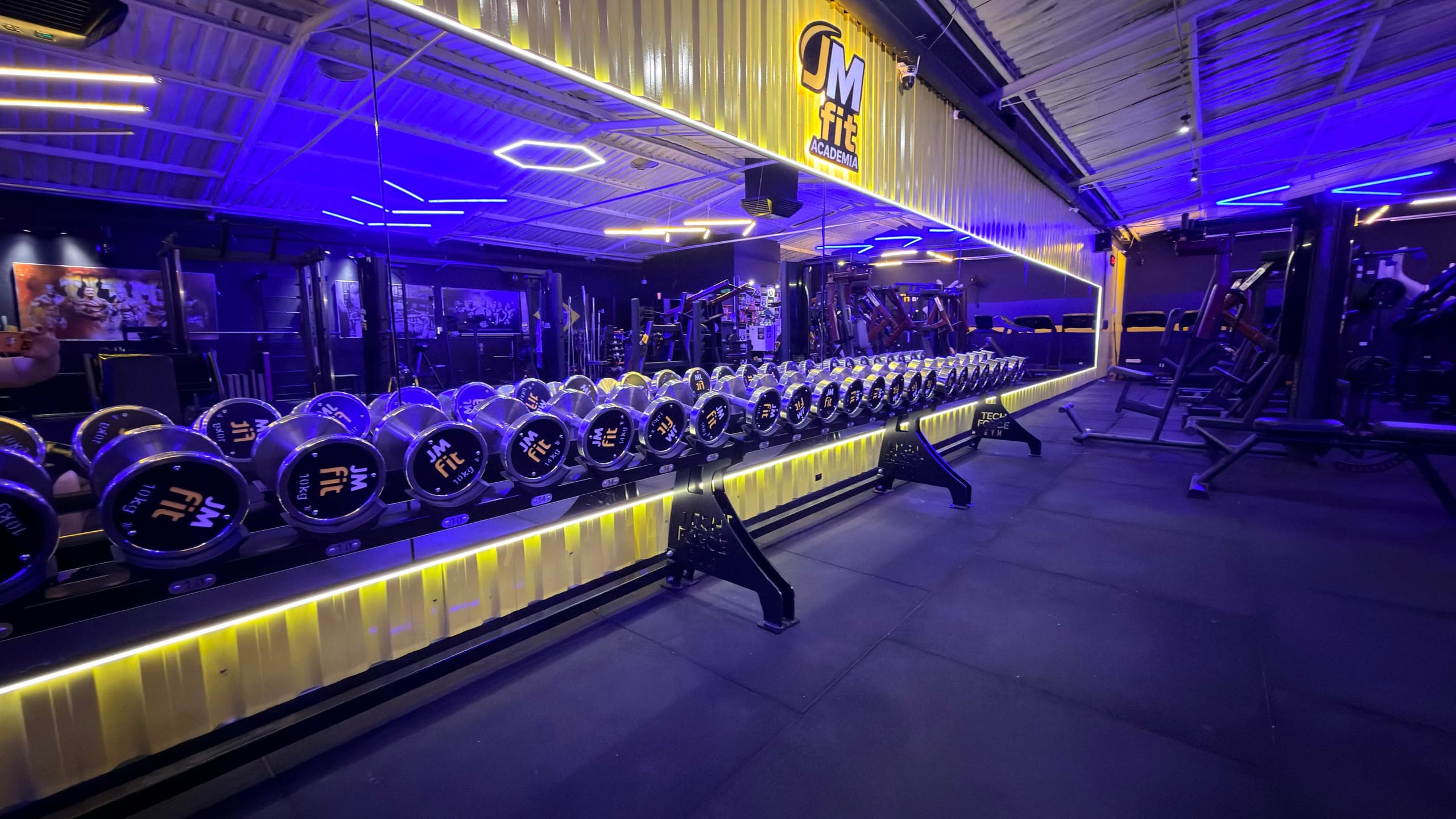 Rack de halteres da JMfit com a marca nos discos e o logo iluminado na parede